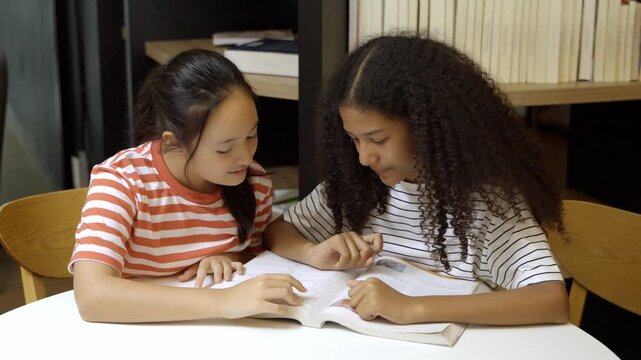 Two girls study together in a library while sharing a book and discussing their schoolwork