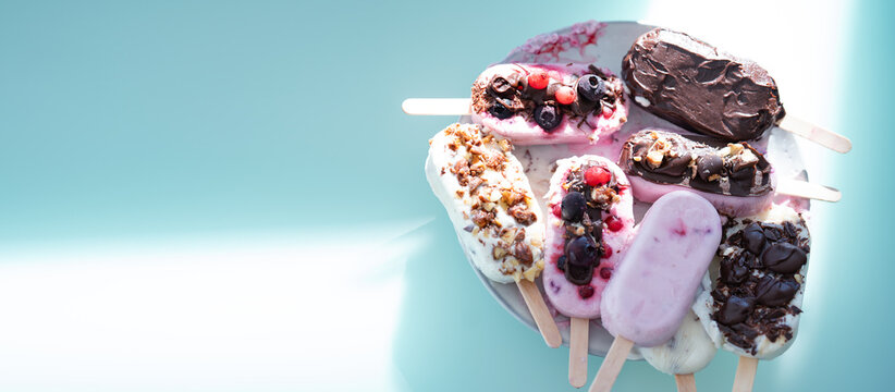 Colorful assortment of homemade yogurt popsicles. Vanilla and chocolate decorated with nuts and frozen berries on ice blue background. Perfect for summer children's birthday party. Vertical overhead c