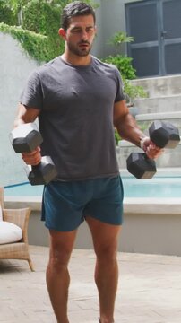 Vertical video: Lowering dumbbells, man in dark shirt and teal shorts curling for strength by pool