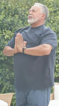 Vertical video: Breathing senior man pressing palms tilting chin up on patio, in navy tee and watch