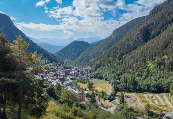 Elevated view of Arinsal village nestled in a narrow Pyrenean valley in Andorra, with traditional stone buildings, dense pine forest, mountain bike track and layered ridges under a blue cloudy sky © IgorsPerspective