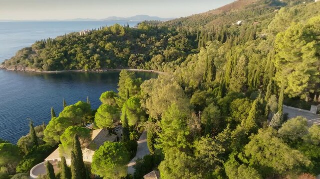 Luxury Mediterranean villas sit on green rocky coast of Corfu island, Greece. UHD aerial explores lush pine trees and exclusive coastal architecture bordering calm sea in Corfu