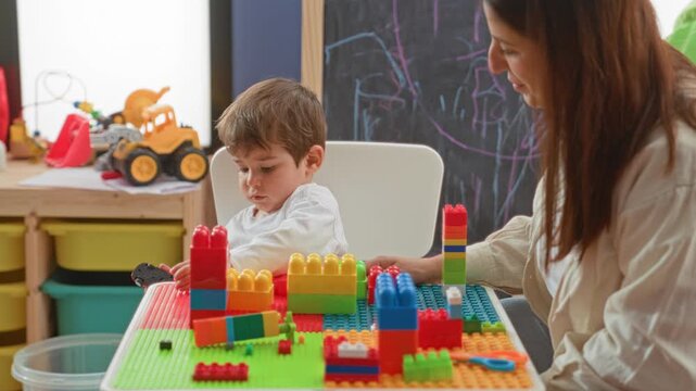 Woman guiding little boy's hand to colorful building blocks on play table near chalkboard in classroom; learning patience.