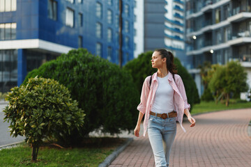 Fototapeta premium Young woman strolls along a sunny city street in jeans and a pink plaid shirt, radiating confident student energy as she walks past glass towers and green spaces.