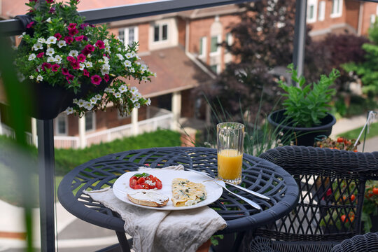 Eye-level wide shot showing breakfast plate with omelet and orange juice on black metal table with hanging flower basket and blurred brick house background