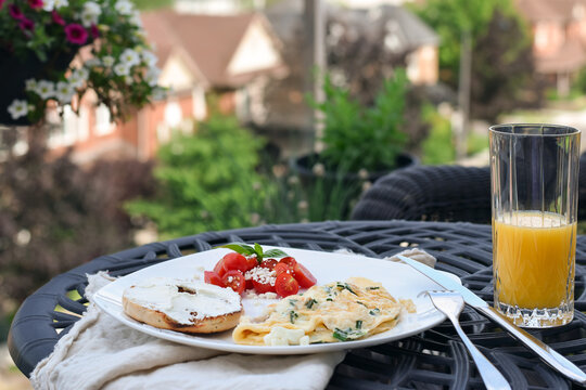 Eye-level close-up showing breakfast plate with omelet, bagel, tomato salad and glass of orange juice on black metal table with blurred neighborhood background