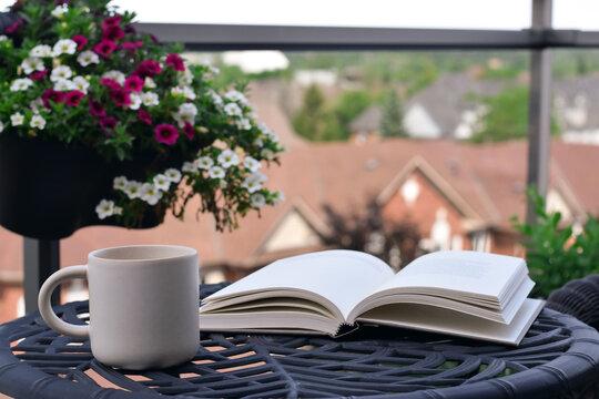 Eye-level close-up showing open book and white mug on black metal table with hanging flower basket and blurred rooftop background