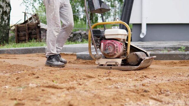 Low angle construction scene shows worker legs and boots moving past plate compactor on sandy surface at outdoor jobsite, with stacked blocks, trees, blurred background, and