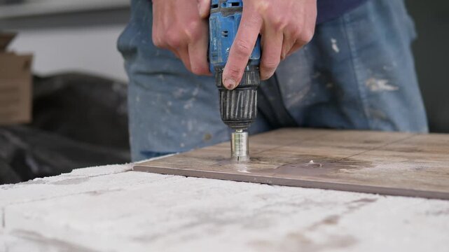 Hands hold blue cordless drill vertically over wood plank on dusty work surface in softly blurred workshop, centered close view with natural light, wood grain, metal chuck, and
