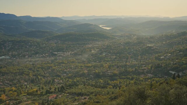 Panoramic cinematic view over the green valley of Pays de Grasse from Cabris, France. Features traditional villas, rolling hills, and Lake Saint-Cassien shimmering under a hazy afternoon sun in Europe