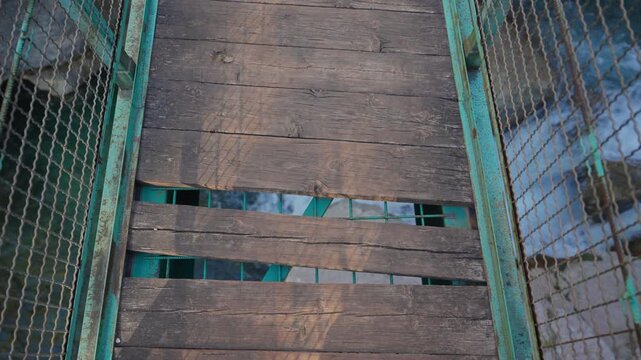 Point of view shot walking across a dangerous and unsafe rickety wooden bridge with missing planks, revealing the rushing river water flowing rapidly underneath the dilapidated structure