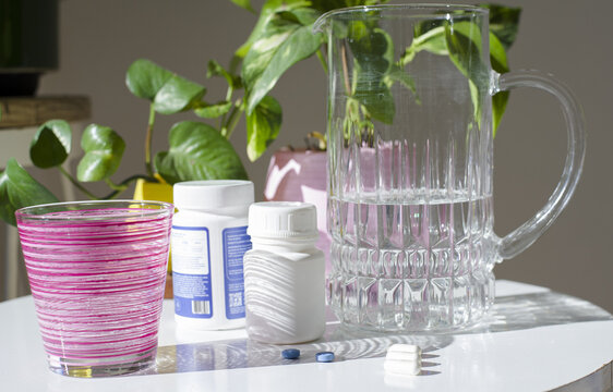 Medicamentos y rutina de salud: pildoras azules y capsulas blancas sobre mesa blanca junTo a frascos, jarra con agua, vaso y una planta