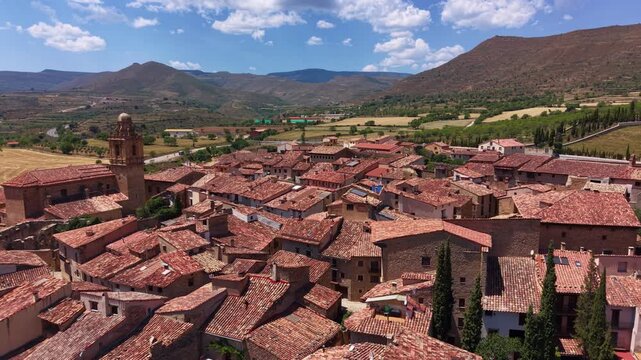 Overhead view of Mirambel, a preserved medieval village in Maestrazgo, Teruel, Spain, featuring terracotta rooftops, a stone bell tower, and surrounding harvested fields and mountain ridges