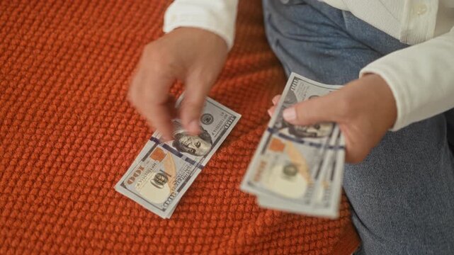 Woman counting stacks of hundred dollar bills with hands on an orange textured bedspread on bed in building; focused saving.