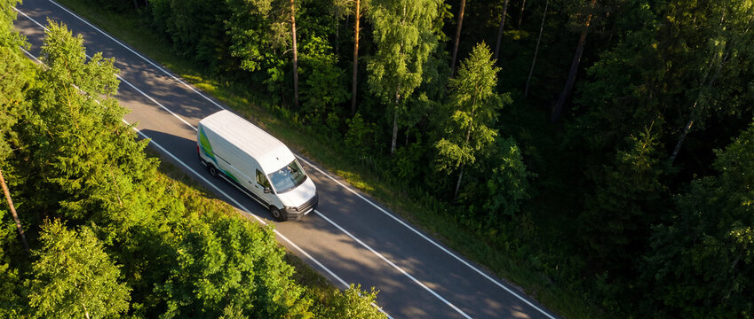 Aerial view white EV van forest road driving along a sunlit two-lane asphalt highway surrounded by dense green summer trees, illustrating sustainable eco-friendly commercial delivery logistics
