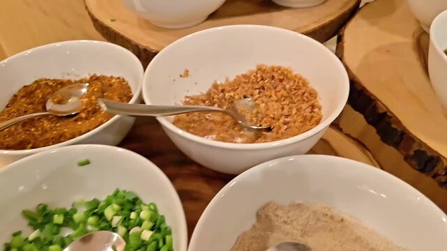 Bowls of chili pepper powder, chopped, peppercorn, and garlic chip condiments with spoons, Food Flavor Garnishing, Tasty Ingredients, Buffet Feast Service, Panning Shot, Slow Motion