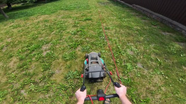 First person view guiding electric corded lawn mower across patchy backyard grass with visible cut swath, trailing power cord, and hands on handlebars during outdoor yard care