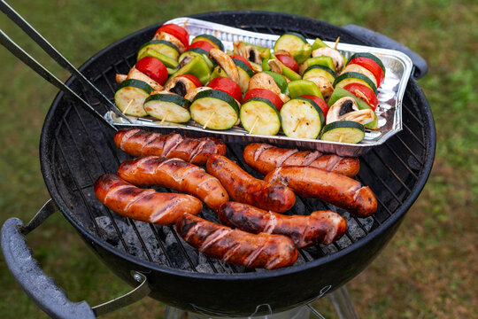 Grilled sausages and vegetable skewers on barbecue. Grilled sausages cook on a charcoal barbecue beside colorful vegetable skewers with zucchini, peppers, mushrooms and tomatoes.