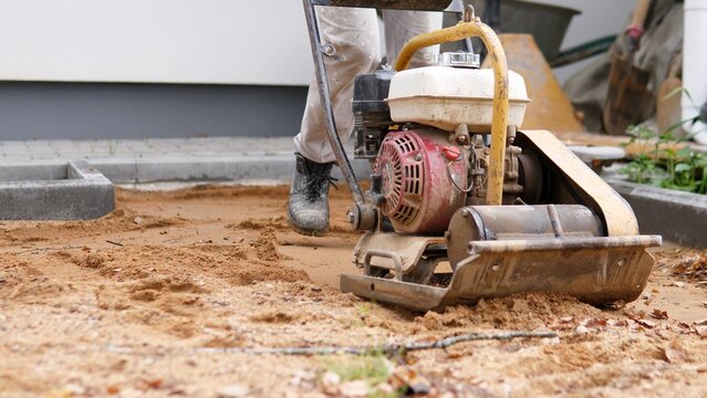 Plate compactor working on sandy ground beside concrete curb during outdoor construction site preparation, with worker boots, leveling base, and unfinished paving edges in soft
