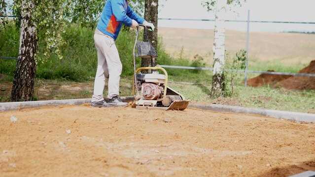 Worker in blue jacket operates vibrating plate compactor on level sand and soil beside curbing, birch trees, fence rails, and open countryside under bright daylight, with worker