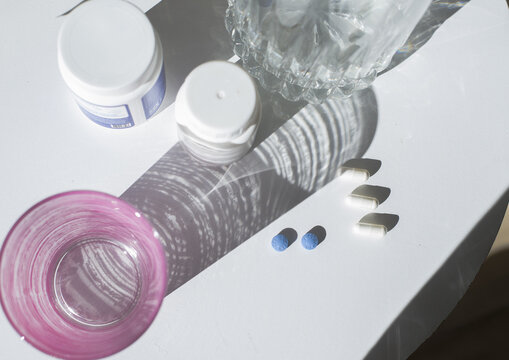Flat lay de Medicamentos y rutina de salud: pildoras azules y capsulas blancas sobre superficie blanca junTo a frascos y jarra con agua