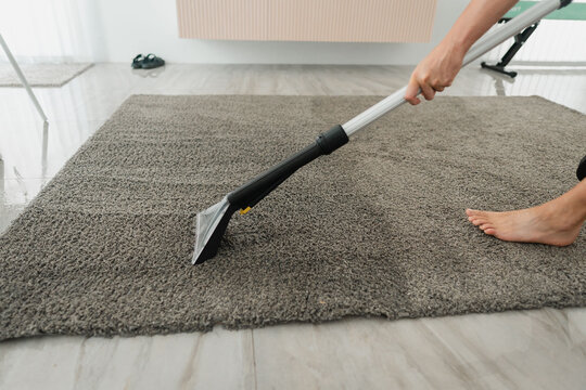 Handheld upholstery tool cleaning gray shag rug on tiled floor beside radiator with person holding wand and bare foot visible in bright natural light, with rug, carpet, cleaning, upholstery detail