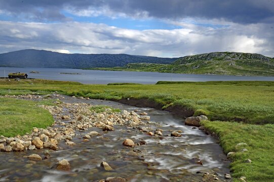 A small river flows through green fields under a cloudy sky with mountains in the background, Fell, Jotumheimen, Norway