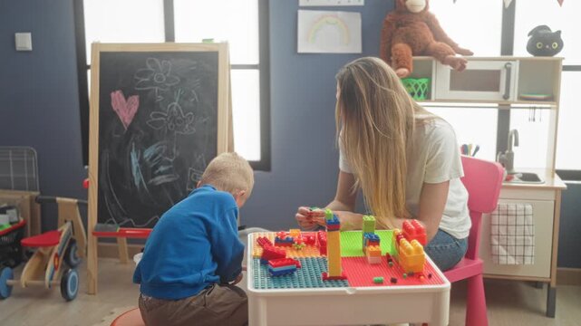 Woman teacher assembles colorful toy blocks with boy at a playtable beside a chalkboard in building; playful learning.