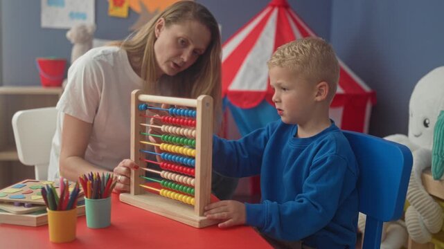 Woman guides boy moving beads on wooden abacus at red activity table in preschool classroom; early learning guidance calm.