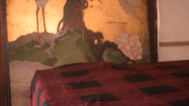 Picking up an old, brown teddy bear and its hat from the foot of a bed covered with a quilt laden with a red and black pattern inside a sunny bedroom