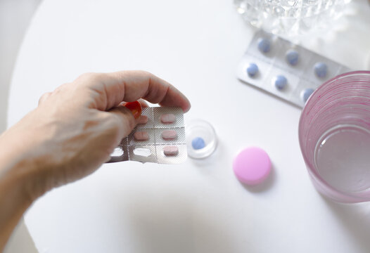 Mano sosteniendo bl&iacute;ster de pastillas rosas sobre mesa blanca con vaso de agua, un pastillaro y pastillas azules