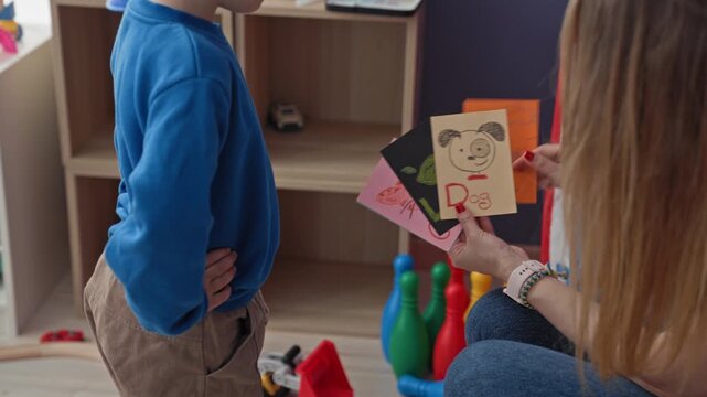 Woman points finger at illustrated flashcards while a boy stands hands on hips beside colorful toy pins and play tent in a playroom; playful learning.