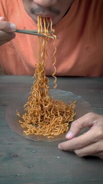 Man eating instant fried noodles with chopsticks at home, close-up comfort food concept, hungry expression, casual lifestyle.