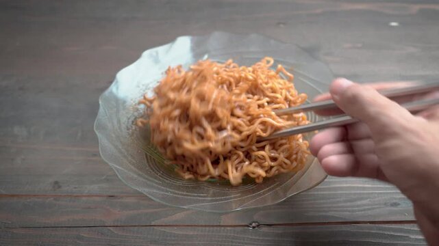 Hand holding chopsticks lifting fried instant noodles on a plate, close-up homemade meal on a wooden table with copy space.