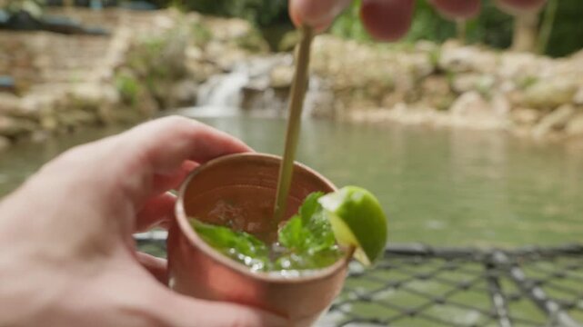A person holds a copper mug filled with a Moscow Mule cocktail garnished with a lime wedge and mint leaves set against a scenic backdrop of a river and lush greenery.