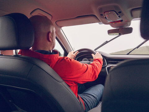 Man drives car, hands on steering wheel. Red jacket, bald head visible from back seat. Interior view shows dashboard and windshield. Daylight outside, road ahead. No animals or plants present