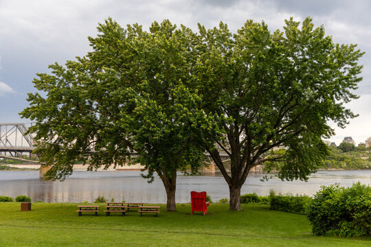 Waterfront park with picnic tables next to Alexandra Bridge in Gatineau. Red Adirondack chair under green trees by river during summer.