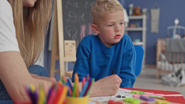 Boy holds marker and draws on paper while woman guides, hand pointing to shapes, blackboard and toys visible inside a building; concentration learning.
