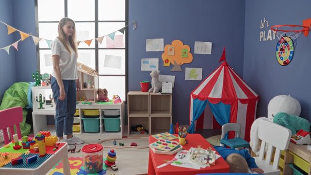 Woman holding toy car points to boy at cluttered red table in playroom building; parental care tenderness.