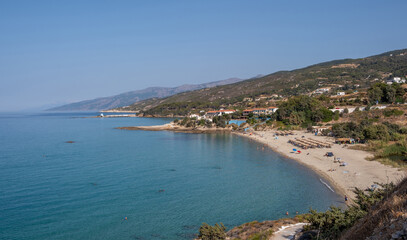 Naklejka premium Mesakti sandy beach and calm turquoise bay on Ikaria Island, Greece. Swimmers and hillside homes, sunny summer day