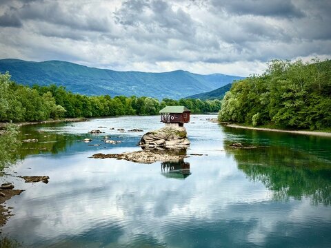 A small wooden house sits atop a rock in the middle of the Drina River in Serbia. Surrounded by lush green forests and mountainous terrain, the house appears isolated and tranquil. The water is calm