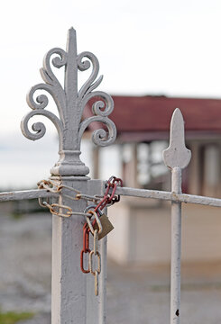 Copper padlock and old iron red and white chain with rusty shackles closing ornate pale painted metal fence. Selective focus