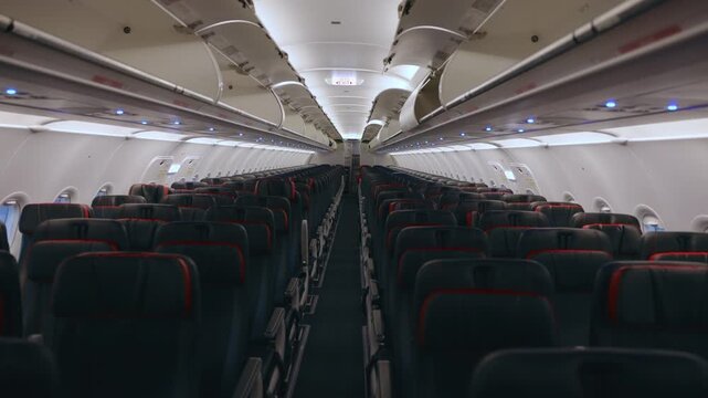 Empty commercial airplane cabin interior featuring rows of dark passenger seats with red trim accents. Overhead compartments and exit signs line the aisle under soft ambient lighting.