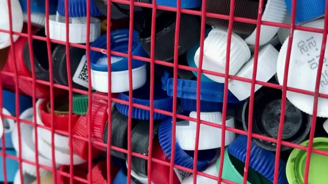 Colorful plastic bottle caps are visibly collected in a large red metal mesh container outdoors