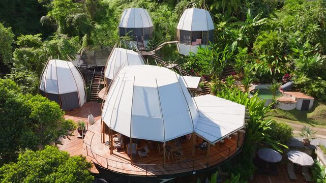 Aerial drone footage captures futuristic pod-shaped glamping resort nestled in lush tropical rainforest with white tent structures, wooden deck platforms, elevated cocoon cabins.