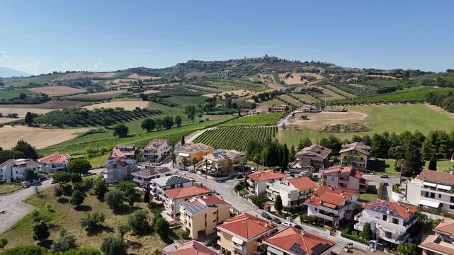 High angle drone view of Roseto degli Abruzzi town residential houses with solar panels near green vineyards and hills under blue sky in Italy.