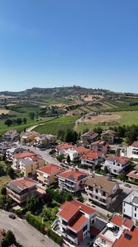 High angle drone view of Roseto degli Abruzzi town residential houses with solar panels near green vineyards and hills under blue sky in Italy.
