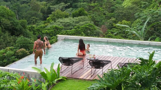 Group of young people dancing by the pool at a small party surrounded by lush tropical jungle. Friends enjoy cocktails on wooden deck with infinity pool overlooking rainforest in slow motion.