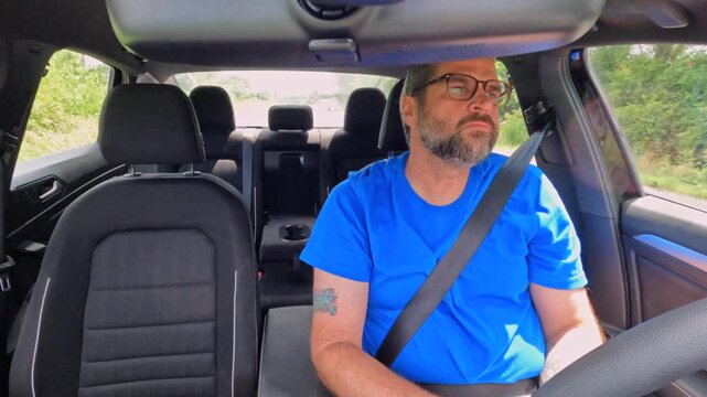Middle aged man wearing blue shirt and glasses driving compact car, looking out the window and stretching his sore neck from long distance driving
