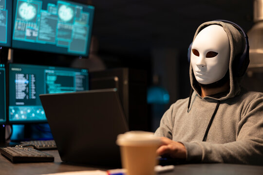 Unidentified man in hoodie wearing white mask as system code displays on screens. Anonymous hacker in office suggests digital secrecy, cybersecurity threats and hidden activity within tech environm.